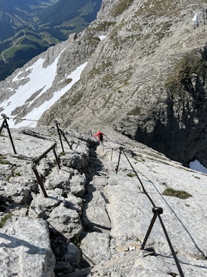 A narrow and steep rocky mountain path is equipped with metal railings for safety, leading up to a high altitude. The path is surrounded by rugged mountain terrain with patches of snow, overlooking a scenic view of a valley with forests and winding roads.