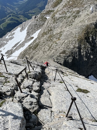 A narrow and steep rocky mountain path is equipped with metal railings for safety, leading up to a high altitude. The path is surrounded by rugged mountain terrain with patches of snow, overlooking a scenic view of a valley with forests and winding roads.