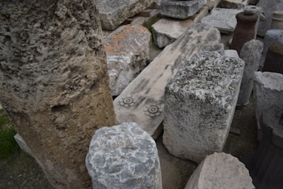 A collection of ancient stone artifacts, including blocks and columns, some with carvings. The stones vary in color and texture, arranged randomly on a concrete surface. Among them, a stone slab features a floral or sun-like design.