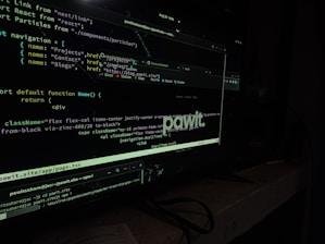 A computer screen displaying code in a dark-themed text editor. The code appears to be written in JavaScript, featuring functions and navigation links. There are file paths and command line interface windows open. A dark environment enhances the glow of the screen.