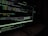 A computer screen displaying code in a dark-themed text editor. The code appears to be written in JavaScript, featuring functions and navigation links. There are file paths and command line interface windows open. A dark environment enhances the glow of the screen.