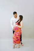 A couple stands together, with the man behind the woman, who is visibly pregnant. The man is dressed in a white shirt and black pants, while the woman wears a vibrant dress with orange and pink patterns. Both display a serene and happy expression.