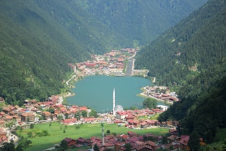 A picturesque village lies nestled between lush green mountains with a serene lake at its center. The village is characterized by its red-roofed buildings and a prominent white mosque with a tall minaret in the foreground. The surrounding dense forest and the blue-green lake create a tranquil and scenic atmosphere.