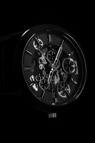 A close-up view of a mechanical watch displaying intricate gears and dials. The watch face is partially illuminated, highlighting the metallic components against a dark background.