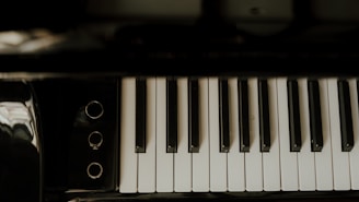 A close-up view of piano keys, highlighting the contrast between the black and white keys. The surface appears polished with a slight reflection visible. Ambient lighting creates a moody and serene atmosphere.