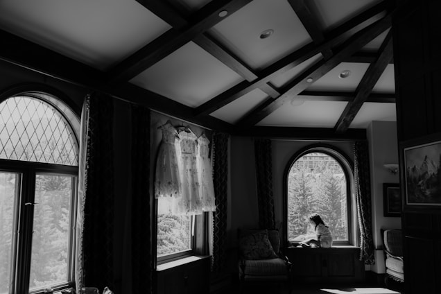 A black and white image of a room with arched windows and wooden beams on the ceiling. Two dresses are hanging by a window, allowing natural light to illuminate them. A girl sits on a window seat, looking outside, creating a contemplative and serene atmosphere.