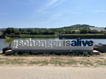 A large sign reading '#schengenisalive' stands in front of a scenic river landscape. The sign is white with blue letters and is surrounded by small plants. In the background, there is a river, greenery, and distant hills under a clear blue sky.