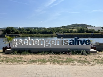 A large sign reading '#schengenisalive' stands in front of a scenic river landscape. The sign is white with blue letters and is surrounded by small plants. In the background, there is a river, greenery, and distant hills under a clear blue sky.
