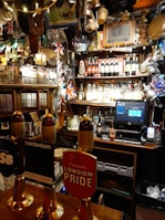 A cozy pub interior with various decorations, including British flags and beer taps prominently displayed. The bar countertop features several beer taps with different labels. Behind the bar, shelves are stocked with a variety of bottles and adorned with unique memorabilia and lights.