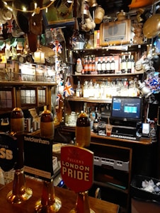 A cozy pub interior with various decorations, including British flags and beer taps prominently displayed. The bar countertop features several beer taps with different labels. Behind the bar, shelves are stocked with a variety of bottles and adorned with unique memorabilia and lights.