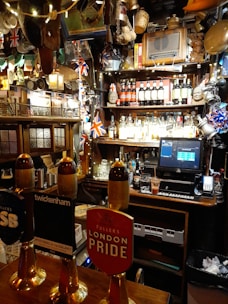 A cozy pub interior with various decorations, including British flags and beer taps prominently displayed. The bar countertop features several beer taps with different labels. Behind the bar, shelves are stocked with a variety of bottles and adorned with unique memorabilia and lights.