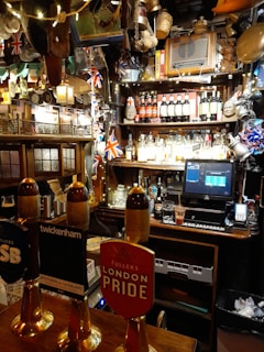 A cozy pub interior with various decorations, including British flags and beer taps prominently displayed. The bar countertop features several beer taps with different labels. Behind the bar, shelves are stocked with a variety of bottles and adorned with unique memorabilia and lights.