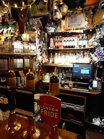 A cozy pub interior with various decorations, including British flags and beer taps prominently displayed. The bar countertop features several beer taps with different labels. Behind the bar, shelves are stocked with a variety of bottles and adorned with unique memorabilia and lights.