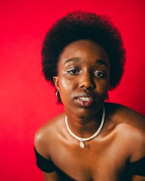 A portrait of a person with short, curly hair and a calm expression against a vibrant red background. They are wearing a necklace with a shell pendant and a black, off-shoulder top.