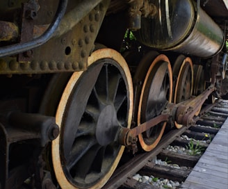Large iron wheels of a vintage steam locomotive are prominently featured, showcasing rust and wear on the metal surfaces. The mechanical components, including thick connecting rods and bolted joints, enhance the industrial feel. Old wooden railway tracks with scattered stones lie beneath, adding to the historical atmosphere.