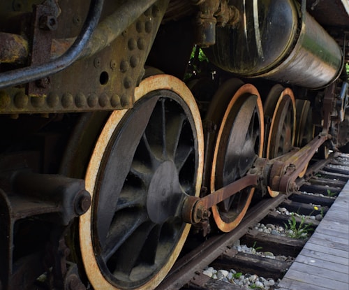 Large iron wheels of a vintage steam locomotive are prominently featured, showcasing rust and wear on the metal surfaces. The mechanical components, including thick connecting rods and bolted joints, enhance the industrial feel. Old wooden railway tracks with scattered stones lie beneath, adding to the historical atmosphere.