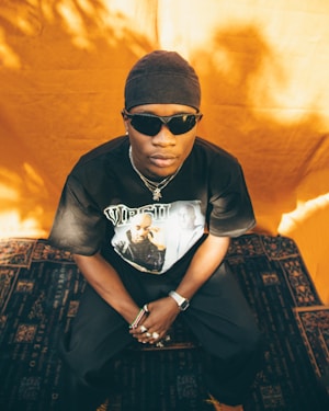 A person wearing a black t-shirt with graphic prints and sunglasses, sitting against an orange backdrop. The individual is accessorized with a watch, rings, and chain necklaces. The surroundings include patterned dark flooring.