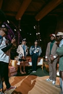 A group of people are gathered indoors, wearing stylish casual and sports-themed outfits, including jackets and caps. Some are standing while one appears to be taking a photo. The environment has a warm, wooden interior with visible beams and spotlights, and a cozy arrangement of furniture including chairs and a sofa.