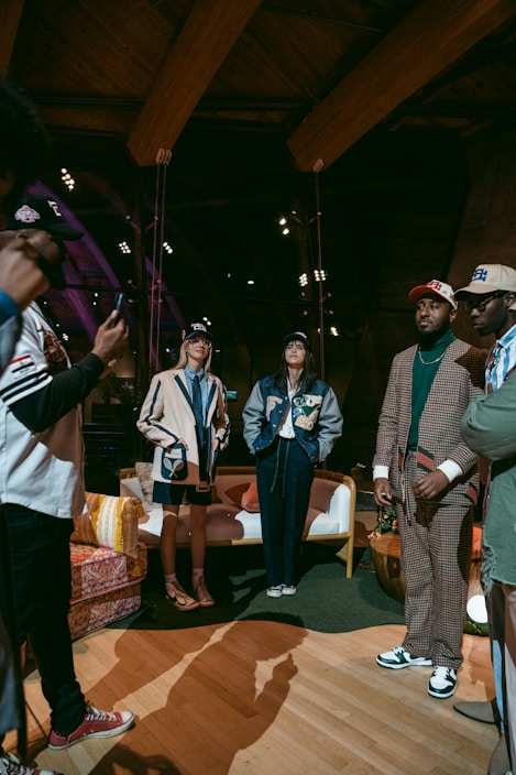 A group of people are gathered indoors, wearing stylish casual and sports-themed outfits, including jackets and caps. Some are standing while one appears to be taking a photo. The environment has a warm, wooden interior with visible beams and spotlights, and a cozy arrangement of furniture including chairs and a sofa.