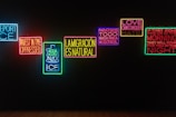 A series of illuminated neon signs are displayed against a dark background. Each sign contains text with messages in different colors, including blue, red, green, yellow, and purple. The phrases express themes of resistance, love, migration, and social justice.