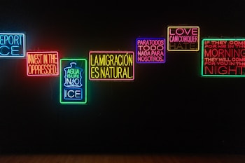 A series of illuminated neon signs are displayed against a dark background. Each sign contains text with messages in different colors, including blue, red, green, yellow, and purple. The phrases express themes of resistance, love, migration, and social justice.