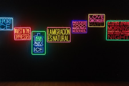 A series of illuminated neon signs are displayed against a dark background. Each sign contains text with messages in different colors, including blue, red, green, yellow, and purple. The phrases express themes of resistance, love, migration, and social justice.