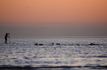A paddleboarder is navigating calm waters during a serene sunset. A group of swimmers are in the water, accompanied by floating markers. The sky exhibits a gentle gradient of orange and pink hues as it transitions into twilight.