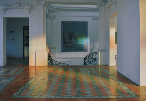 A spacious interior space featuring intricate floor tiles in a checkered pattern with warm tones. The room is adorned with white walls and architectural elements like decorative cornices and pilasters. A muted painting hangs on the wall, and a curved staircase with artistic railing is visible.