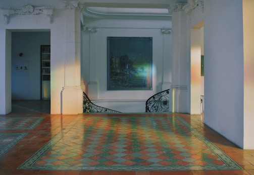 A spacious interior space featuring intricate floor tiles in a checkered pattern with warm tones. The room is adorned with white walls and architectural elements like decorative cornices and pilasters. A muted painting hangs on the wall, and a curved staircase with artistic railing is visible.