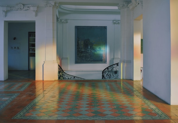 A spacious interior space featuring intricate floor tiles in a checkered pattern with warm tones. The room is adorned with white walls and architectural elements like decorative cornices and pilasters. A muted painting hangs on the wall, and a curved staircase with artistic railing is visible.