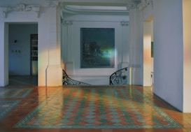 A spacious interior space featuring intricate floor tiles in a checkered pattern with warm tones. The room is adorned with white walls and architectural elements like decorative cornices and pilasters. A muted painting hangs on the wall, and a curved staircase with artistic railing is visible.