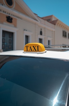 A green and orange taxi with a visible phone number on its sign is parked on a street in front of residential houses. The surrounding area includes trees and a typical suburban neighborhood environment.