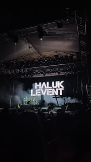 A concert stage is set up with musical instruments like drums and guitars in front of a large screen displaying the name 'HALUK LEVENT'. The lighting creates a dramatic atmosphere with a dark, moody background.