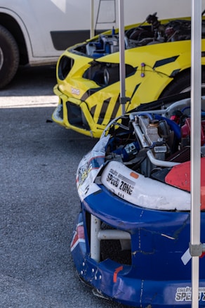 Two race car fronts are prominently displayed. The foreground features a blue and white car with a visible 'SPEED ZONE' decal and parts removed, while in the background, a yellow car with black stripes and exposed engine parts is seen under a tent structure.