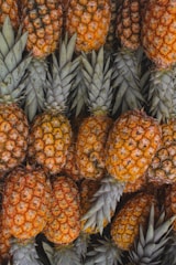 A cluster of ripe pineapples with golden-brown skin and green leaves closely packed together. The texture of the pineapples' spiny exteriors is clearly visible, suggesting freshness.