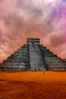 An ancient, large stepped pyramid made of stone, likely a cultural and historical monument. The pyramid has a wide staircase leading to the top. The sky is filled with dramatic, colorful clouds, with hues of purple, pink, and orange. There are people scattered around the base of the pyramid on an orange ground, giving a sense of scale to the massive structure.