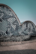 A wavy concrete wall adorned with intricate blue and white mosaic artwork depicting figures and abstract shapes. The mural includes elements like birds and trees, set against a background of leafy patterns. In front, there is a patch of well-kept green grass and a paved stone pathway.