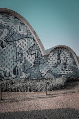 A wavy concrete wall adorned with intricate blue and white mosaic artwork depicting figures and abstract shapes. The mural includes elements like birds and trees, set against a background of leafy patterns. In front, there is a patch of well-kept green grass and a paved stone pathway.