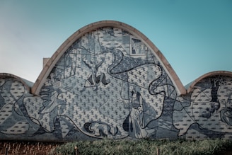 A mural on a curved building wall features abstract and geometric patterns with human figures. The artwork includes intricate designs with people in elaborate poses, surrounded by swirling lines and shapes. The background has repeating motifs, indicating a theme or story.