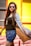 A person wearing sunglasses and casual summer clothing stands against a vibrant, colorful background. They have long hair and are wearing a black top with denim shorts, and a long grey cardigan that billows slightly.