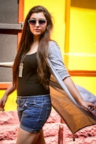A person wearing sunglasses and casual summer clothing stands against a vibrant, colorful background. They have long hair and are wearing a black top with denim shorts, and a long grey cardigan that billows slightly.