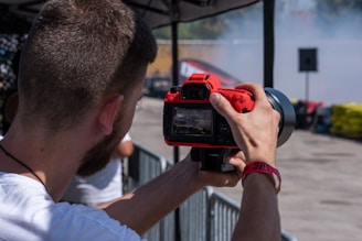 A person is holding a red camera with both hands, capturing a photo or video at an outdoor event. The focus is on the back of the camera and the photographer’s arms, with a blurred background featuring barriers and colorful objects.