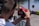 A person is holding a red camera with both hands, capturing a photo or video at an outdoor event. The focus is on the back of the camera and the photographer’s arms, with a blurred background featuring barriers and colorful objects.