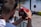 A person is holding a red camera with both hands, capturing a photo or video at an outdoor event. The focus is on the back of the camera and the photographer’s arms, with a blurred background featuring barriers and colorful objects.