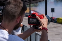 A person is holding a red camera with both hands, capturing a photo or video at an outdoor event. The focus is on the back of the camera and the photographer&rsquo;s arms, with a blurred background featuring barriers and colorful objects.