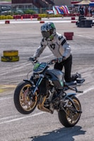 A motorcyclist is performing a wheelie on a powerful, custom-styled bike with a sporty design, wearing full protective gear, including a helmet with a reflective visor and a white jacket. The background features colorful stacked tires and banners, suggesting a closed course or event area, likely for a motorsport or stunt riding competition.