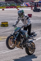 A motorcyclist is performing a wheelie on a powerful, custom-styled bike with a sporty design, wearing full protective gear, including a helmet with a reflective visor and a white jacket. The background features colorful stacked tires and banners, suggesting a closed course or event area, likely for a motorsport or stunt riding competition.