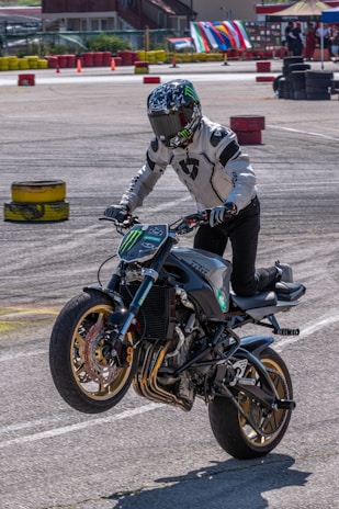 A motorcyclist is performing a wheelie on a powerful, custom-styled bike with a sporty design, wearing full protective gear, including a helmet with a reflective visor and a white jacket. The background features colorful stacked tires and banners, suggesting a closed course or event area, likely for a motorsport or stunt riding competition.