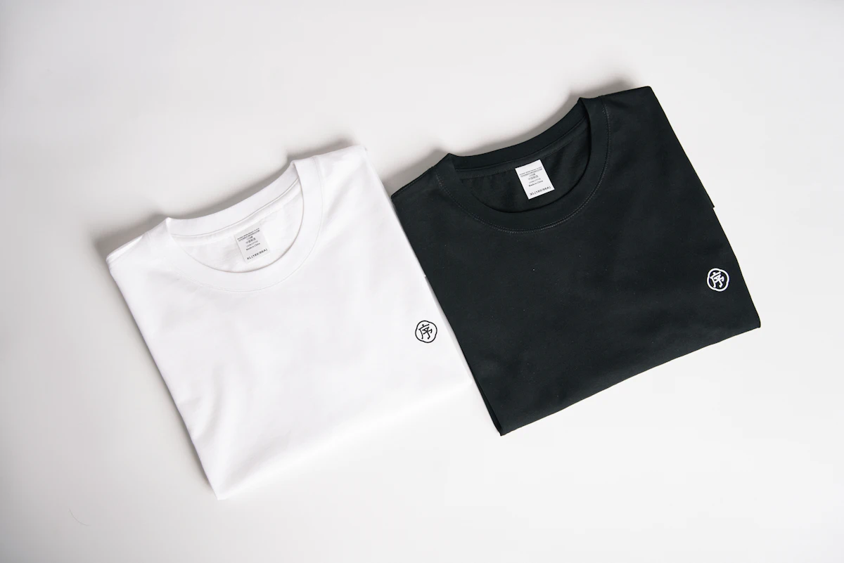 Kaos hitam dan putih dengan sablon logo dada kecil tergeletak rapi sebagai foto produk di latar studio