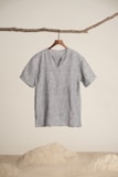 A simplistic short-sleeved, gray linen shirt hangs from a wooden hanger, attached to a natural branch. The background is minimalistic with a sandy floor and white wall.